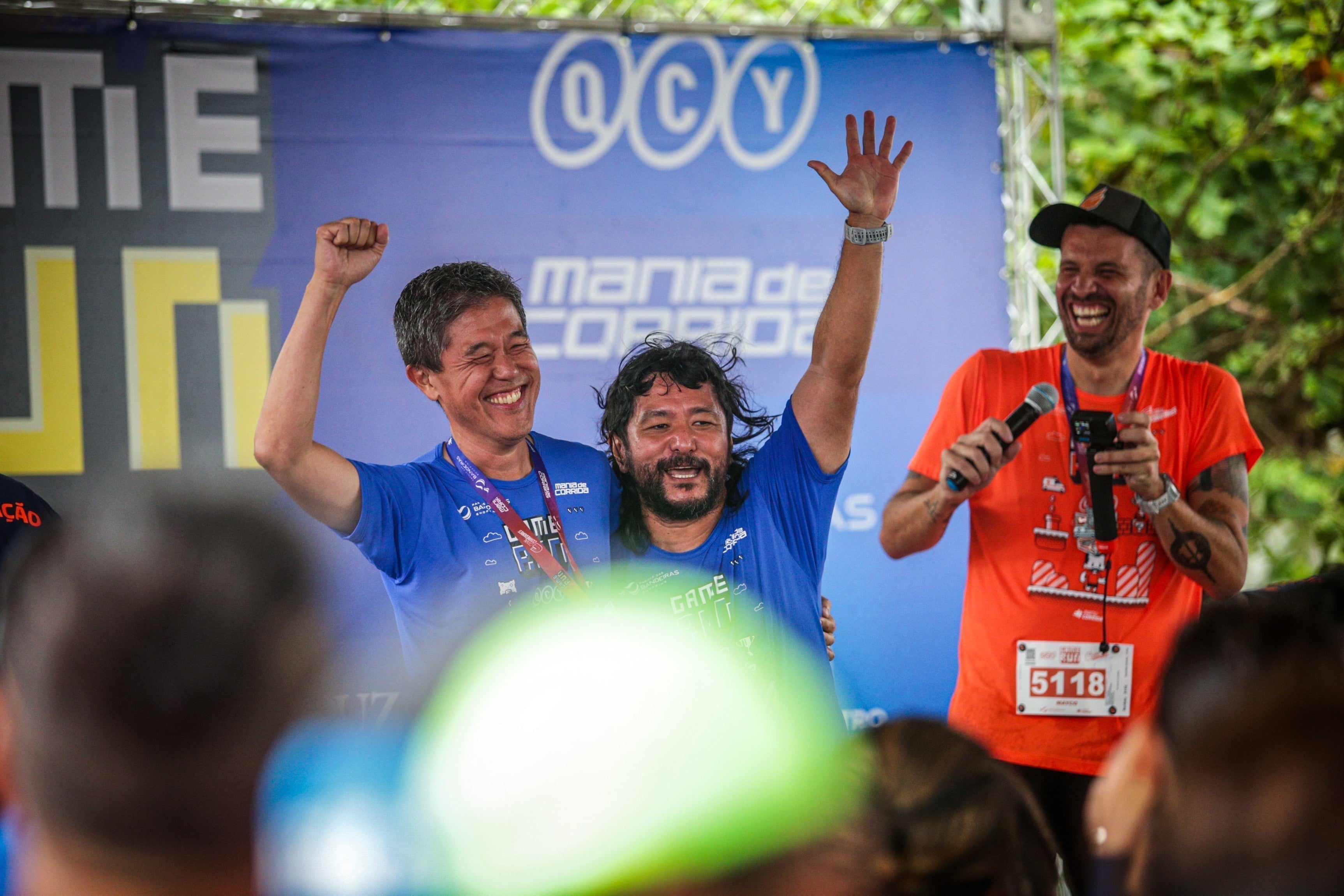 Zwei Läufer jubeln auf dem Podium beim QCY-Laufevent in São Paulo, ein Moderator mit Mikrofon feiert mit.