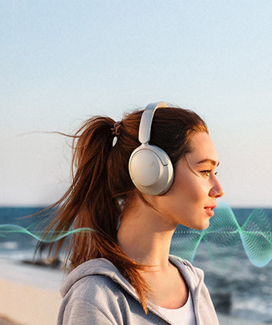 Frau mit weißen Over-Ear-Kopfhörern am Strand, Wind weht durchs Haar.