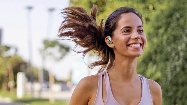 Lächelnde Frau beim Joggen im Park mit kabellosen Kopfhörern, Haare wehen im Wind, sportliches Lifestyle-Foto.