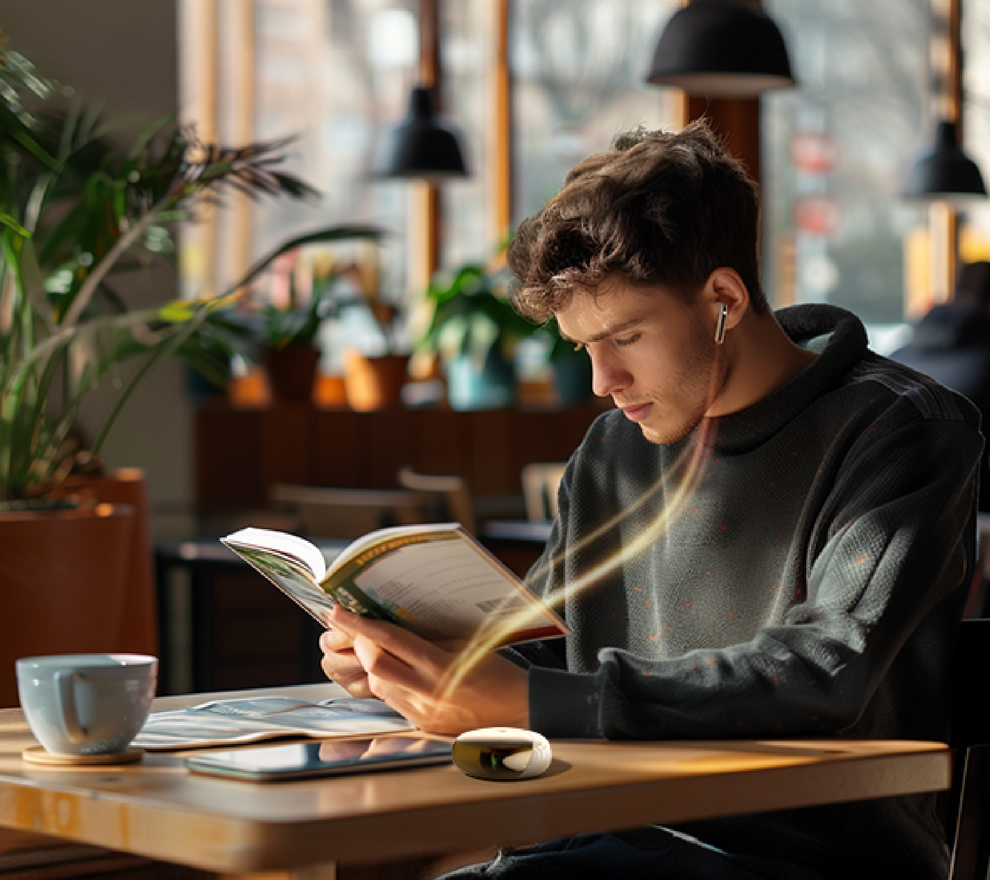 Junger Mann liest Buch im Café mit kabellosen Kopfhörern und Tasse Kaffee auf dem Holztisch.