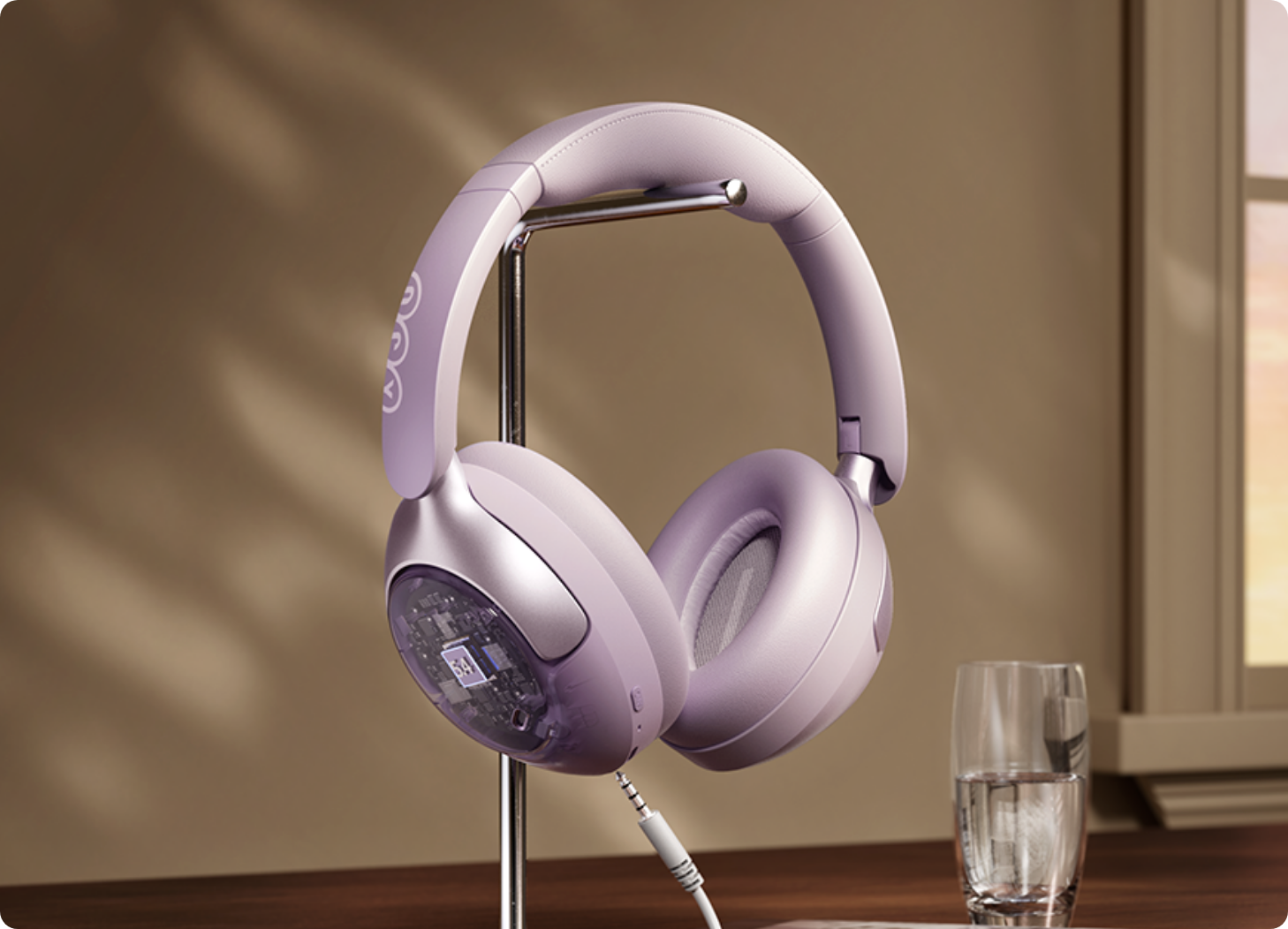 Lavendelfarbene Over-Ear-Kopfhörer mit transparentem Gehäuse auf Ständer, Kabel angeschlossen, neben Wasserglas am Fenster.