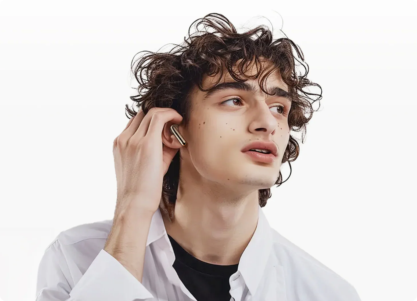 Junger Mann mit lockigem Haar setzt goldene kabellose Ohrhörer ein, weißes Hemd, moderner Lifestyle-Look.