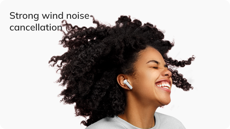 Frau mit lockigem Haar und In-Ear-Kopfhörern, lachend bei starkem Wind – Noise-Cancellation-Funktion wird demonstriert.